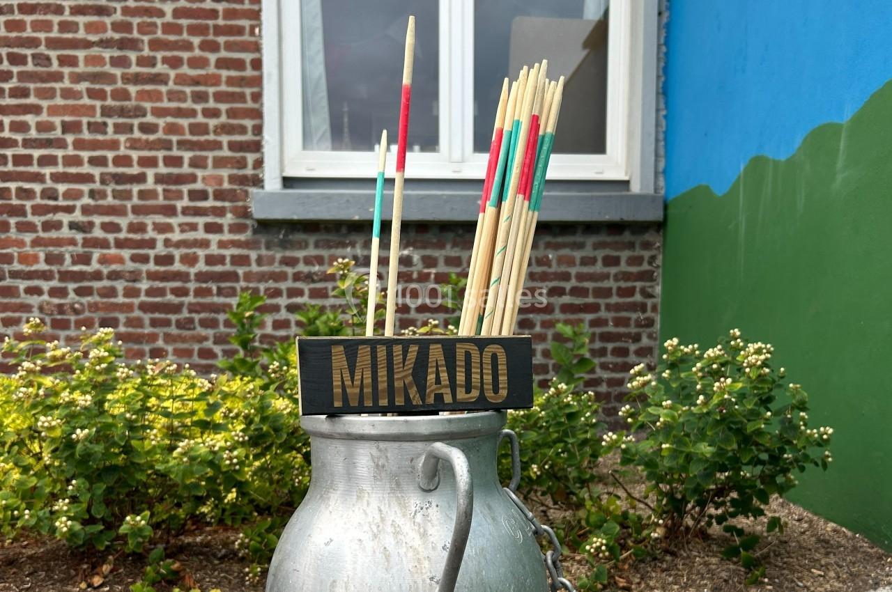 Des bâtons de jeu Mikado colorés placés dans un ancien bidon en métal devant un mur en briques et un jardin.