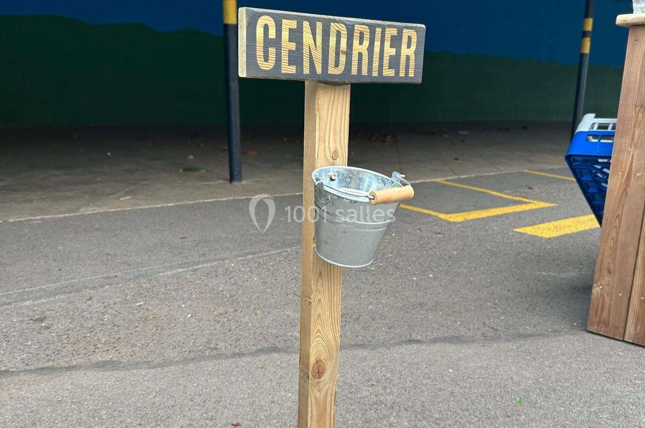 Poteau en bois avec un seau métallique suspendu servant de cendrier, situé sur un sol asphalté.