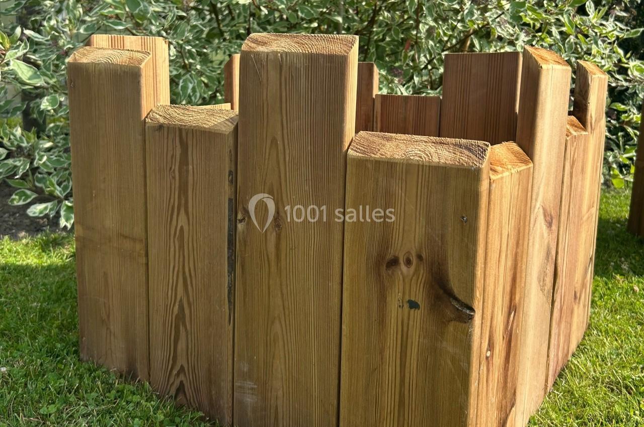 Carré en bois brut assemblé, posé sur une pelouse, entouré de végétation en arrière-plan.