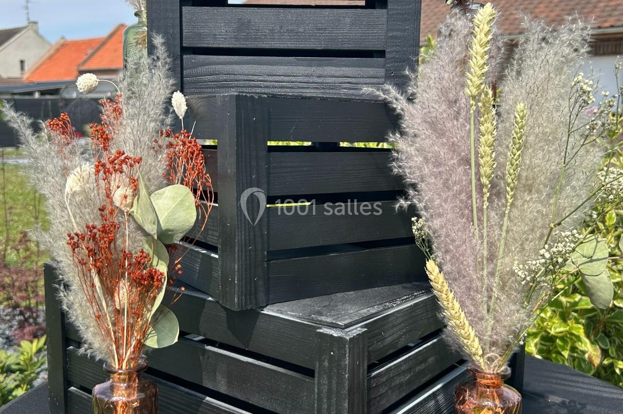 Caisses en bois noires empilées, entourées de bouquets de fleurs séchées dans des vases en verre, en extérieur.