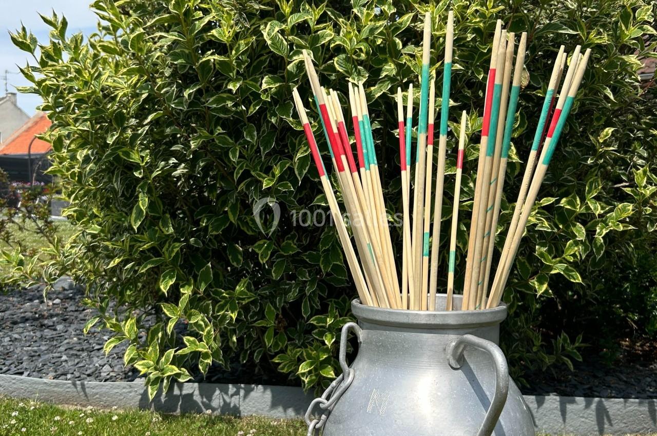 Bâtons de mikado géants colorés dans un pot en métal, posés devant un buisson dans un jardin.