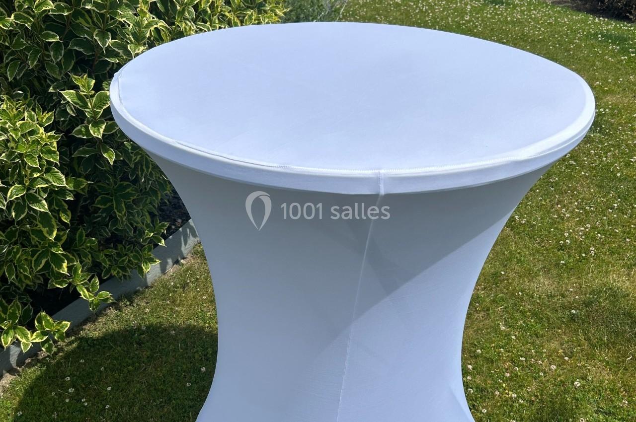 Table haute ronde recouverte d'une housse blanche, placée sur une pelouse près d'arbustes.