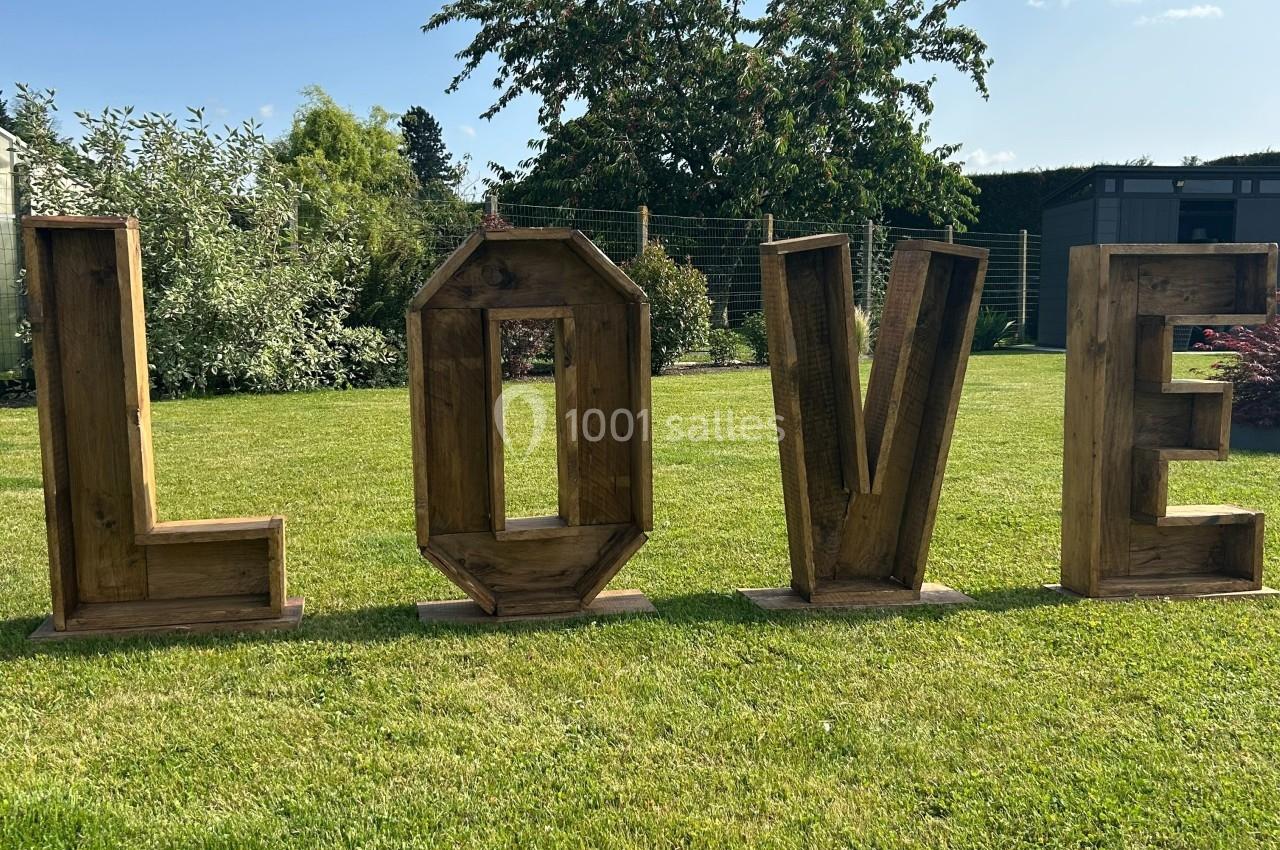 Lettres en bois formant le mot ’LOVE’ disposées sur une pelouse, avec des arbres et un jardin en arrière-plan.