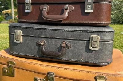 Trois valises vintage empilées, en cuir et tissu, posées sur une surface en extérieur avec de l'herbe en arrière-plan.