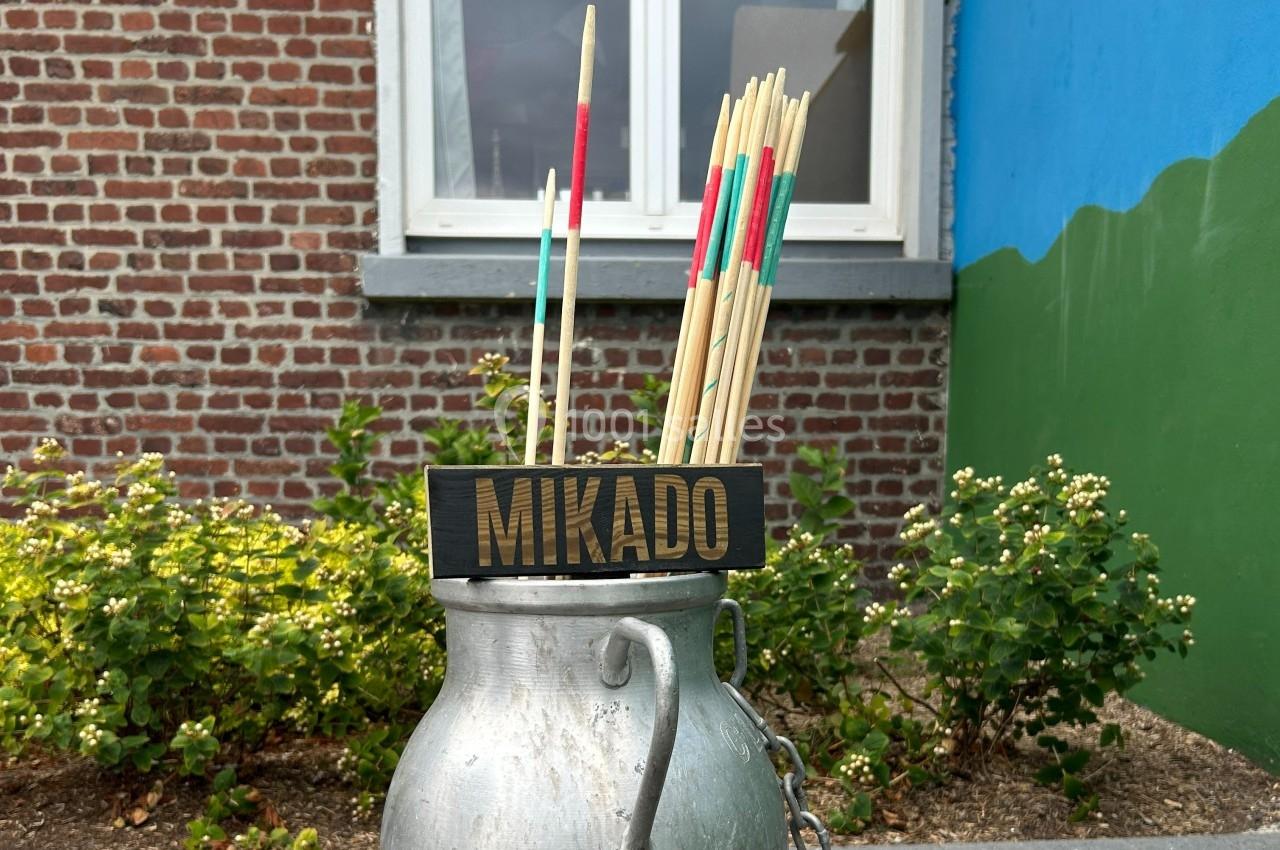 Des bâtons de mikado géants dans un pot en métal devant un mur de briques et un jardin fleuri.