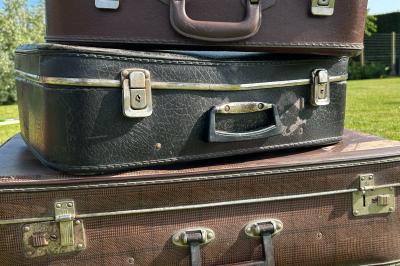 Trois valises vintage empilées, en cuir et tissu, posées sur une surface en extérieur avec de l'herbe en arrière-plan.