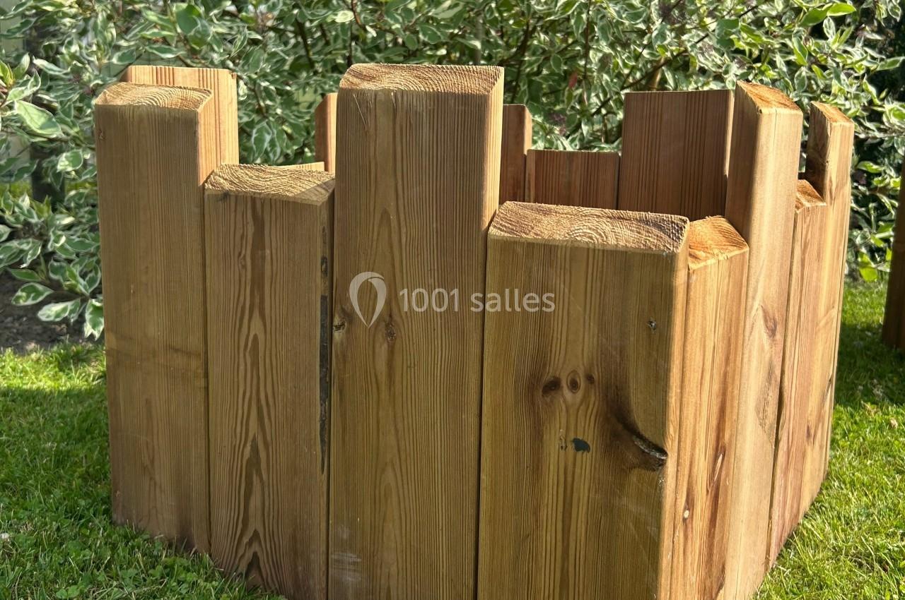 Structure en bois brut formant un cercle, posée sur une pelouse, avec des arbustes en arrière-plan.