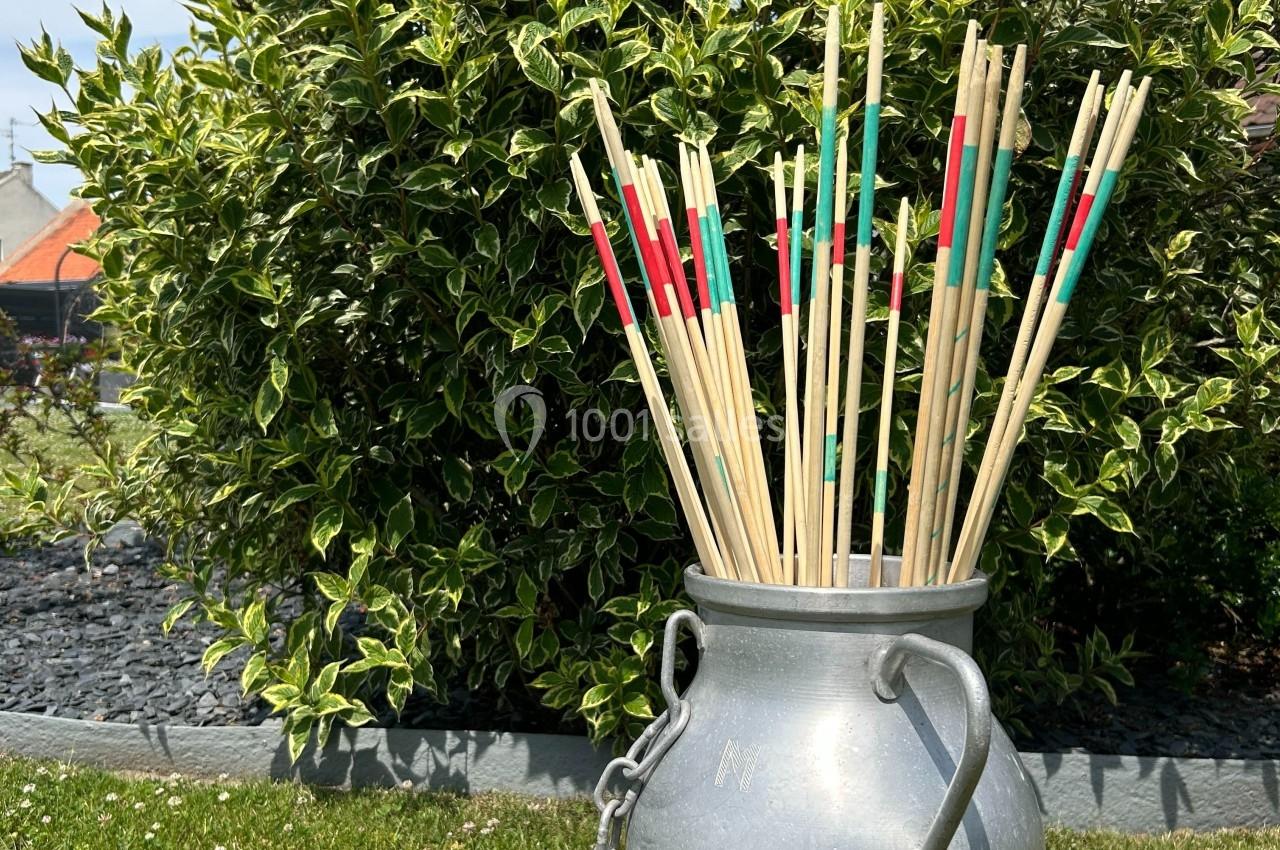 Bâtonnets de mikado géants colorés dans un pot en métal, posés devant un buisson dans un jardin.