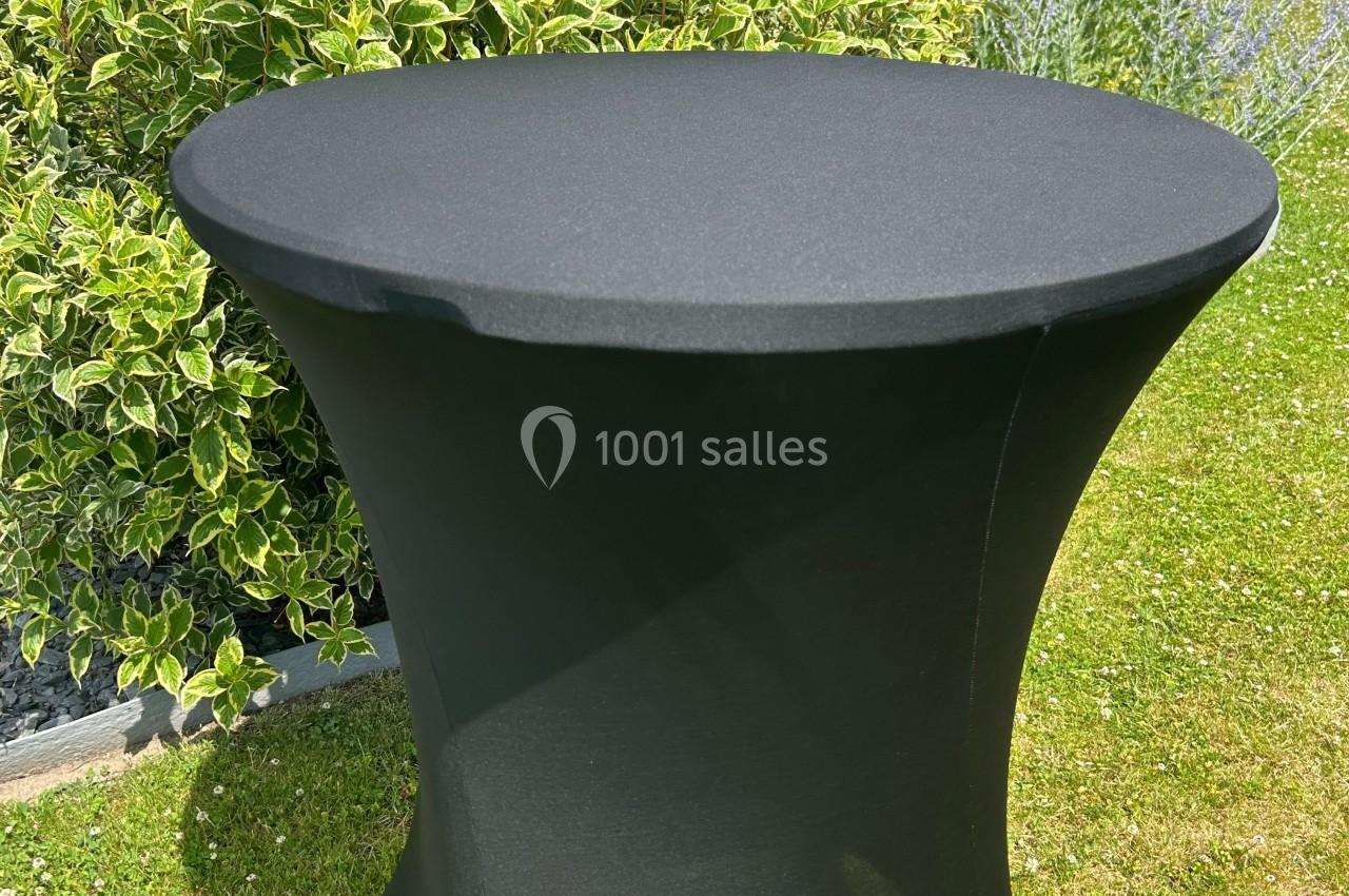 Table ronde noire avec housse ajustée, placée sur une pelouse, entourée de végétation.