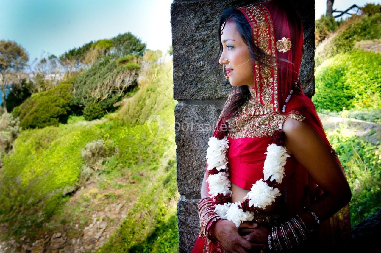 Femme en tenue traditionnelle rouge et dorée, ornée de guirlandes de fleurs blanches, regardant un paysage verdoyant.