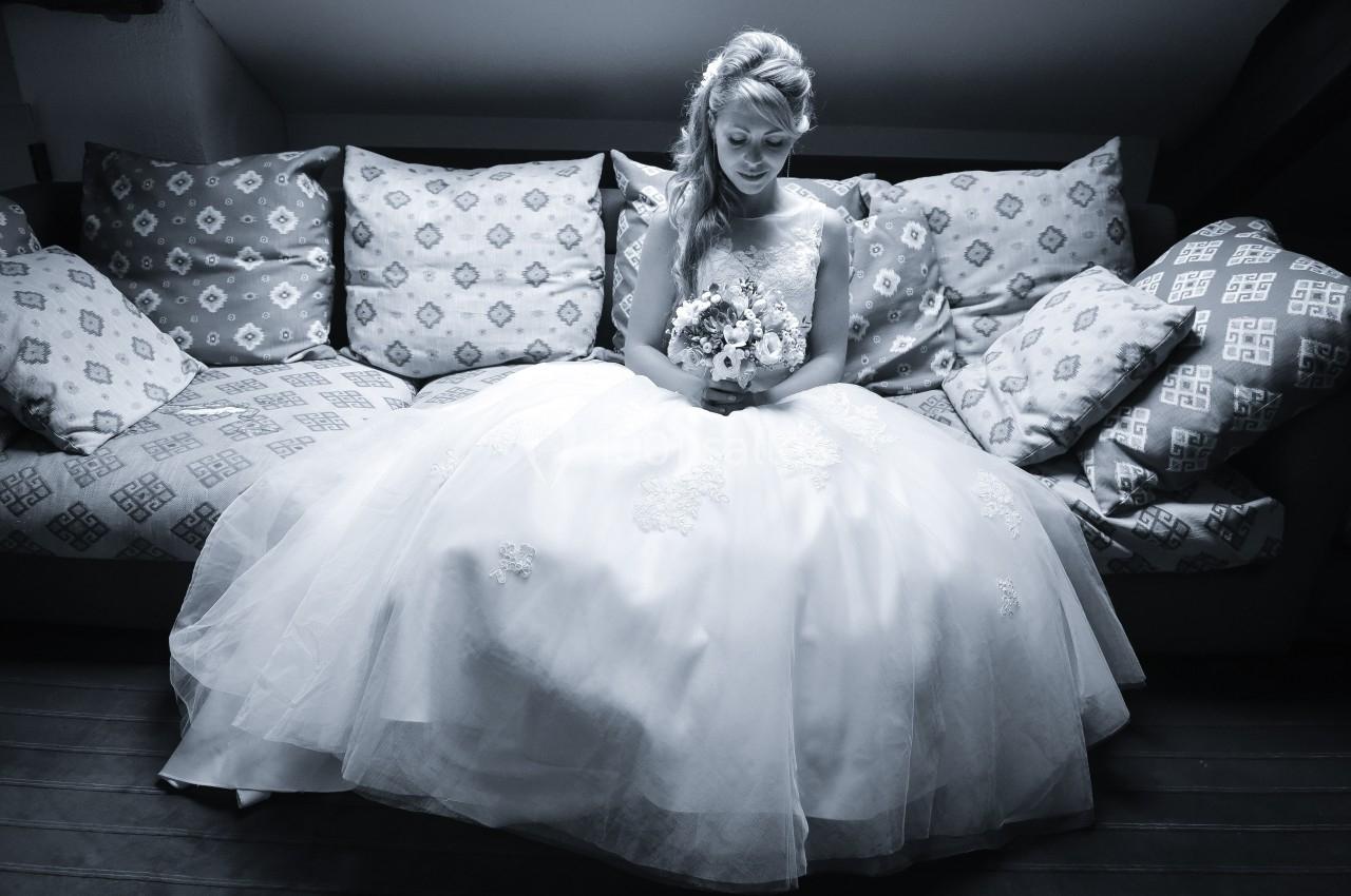 Une femme en robe de mariée assise sur un canapé, tenant un bouquet de fleurs, dans une ambiance tamisée.