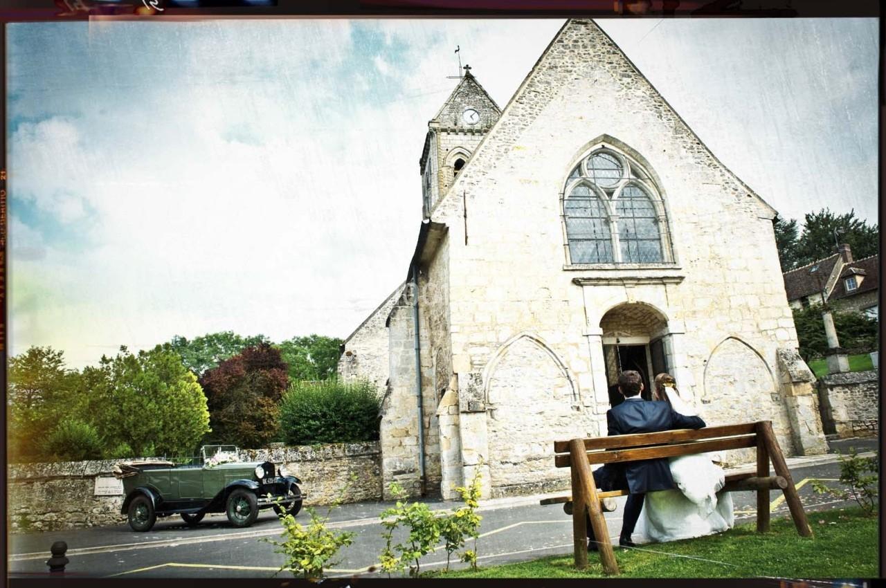 Un couple assis sur un banc devant une église en pierre, avec une voiture ancienne garée à proximité.