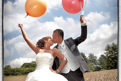 Miniature Victor Podgorski Photographe #15 Un couple en tenue de mariage s'embrasse dans un champ de blé, tenant des ballons colorés sous un ciel bleu.
