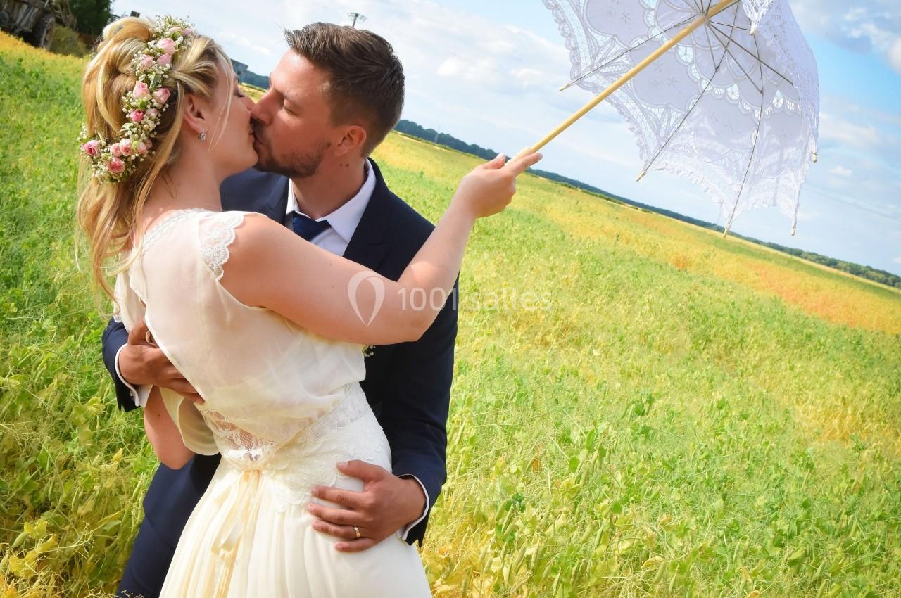 Un couple en tenue de mariage s'embrasse dans un champ sous un ciel bleu, la mariée tenant une ombrelle blanche.