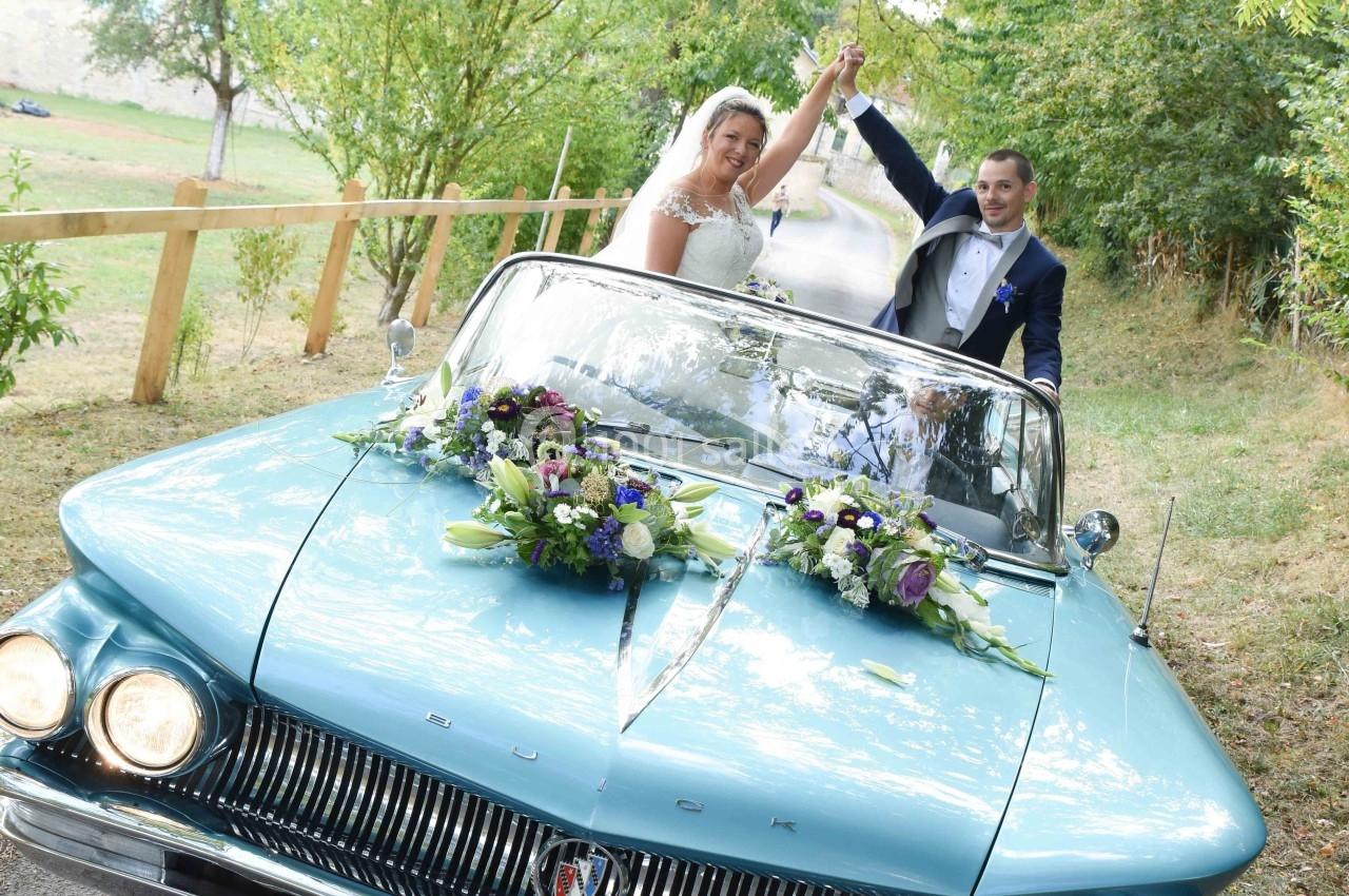 Un couple de mariés souriants dans une voiture décapotable bleue décorée de fleurs, sur une route bordée d'arbres.