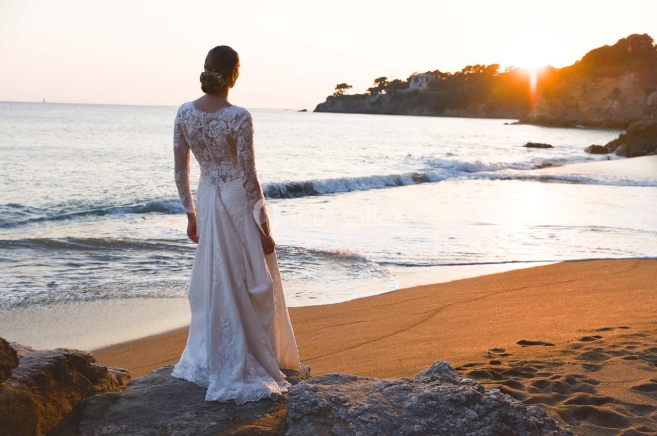 Femme en robe de mariée regardant un coucher de soleil sur une plage, avec des vagues et des rochers au premier plan.