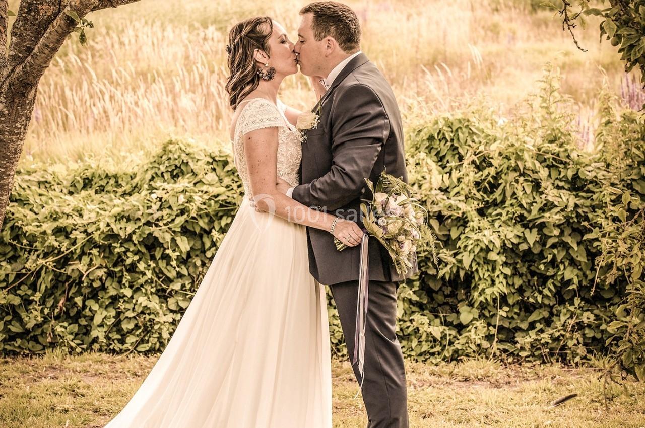 Un couple en tenue de mariage s'embrasse sous un arbre dans un cadre champêtre.