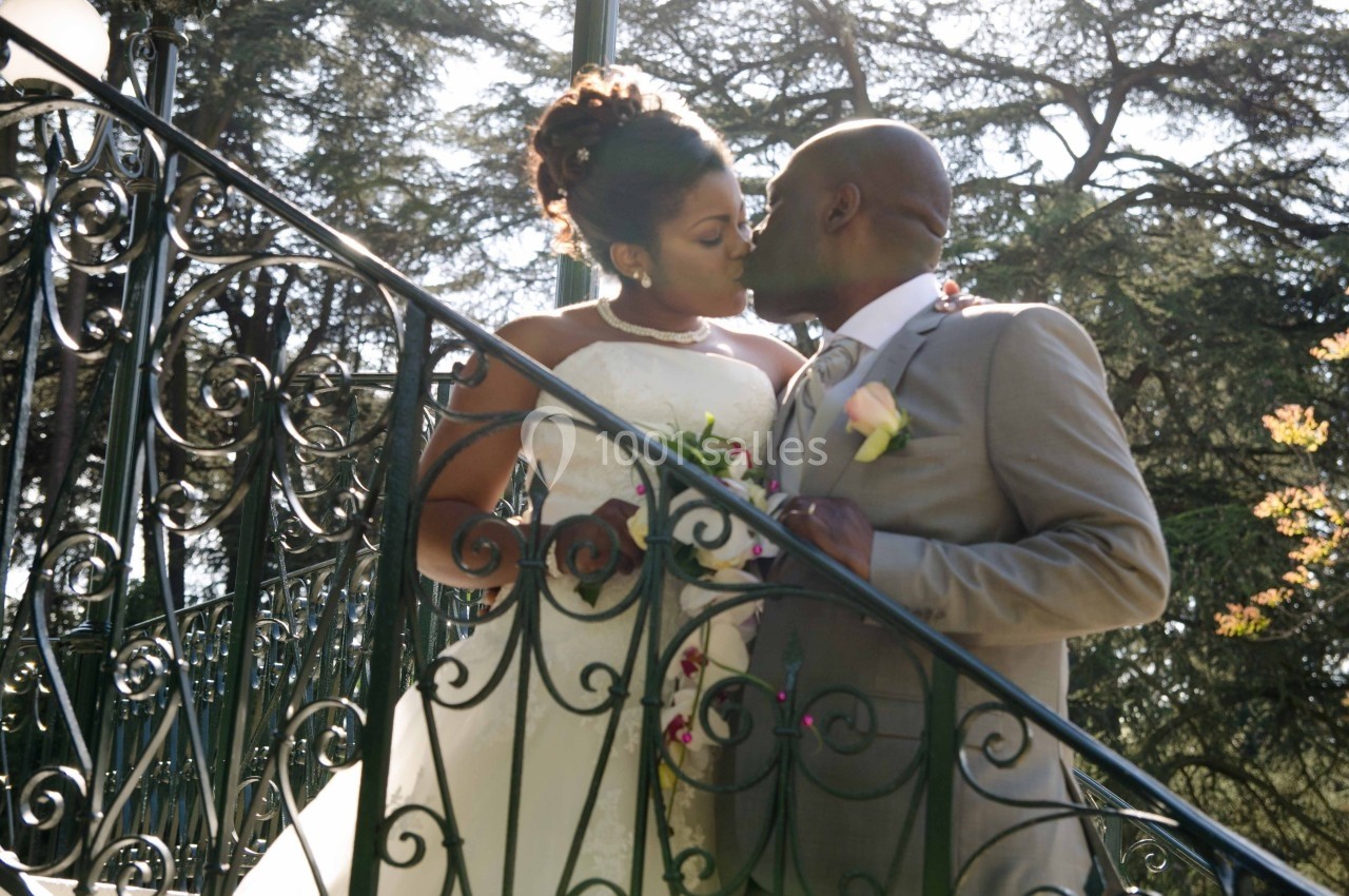 Un couple en tenue de mariage s'embrasse sur un escalier en fer forgé, entouré d'arbres et de lumière naturelle.
