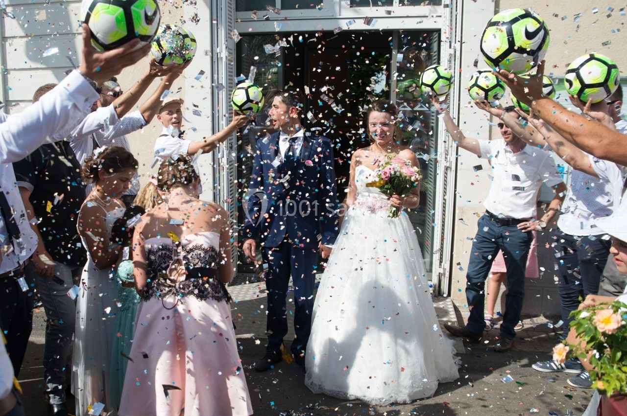 Un couple de mariés sort d'un bâtiment sous une pluie de confettis et des invités tenant des ballons de football.