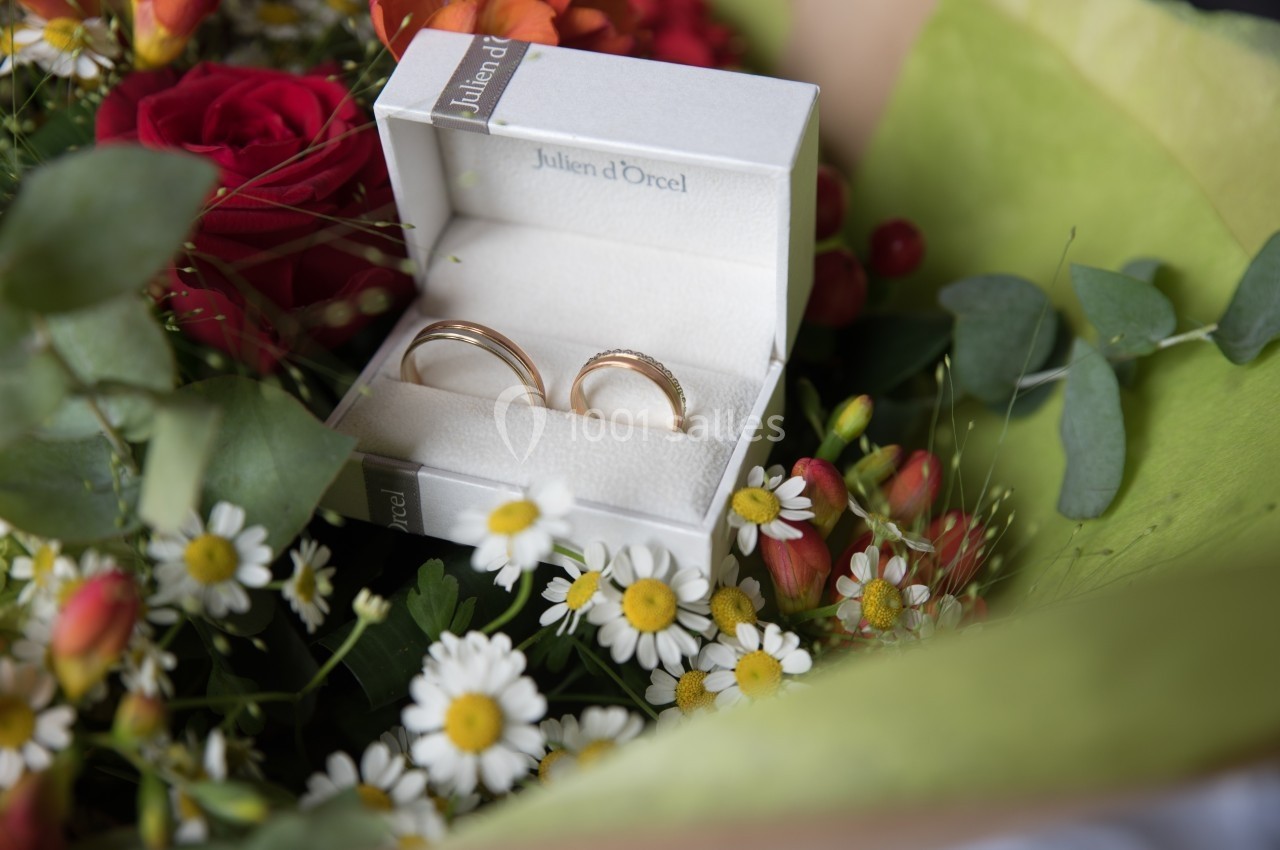 Deux alliances en or dans un écrin blanc, entourées de fleurs colorées et de feuillage.