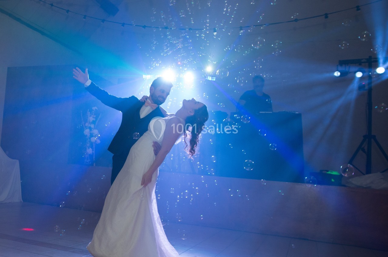 Un couple en tenue de mariage danse sous des bulles et des lumières colorées dans une salle décorée.