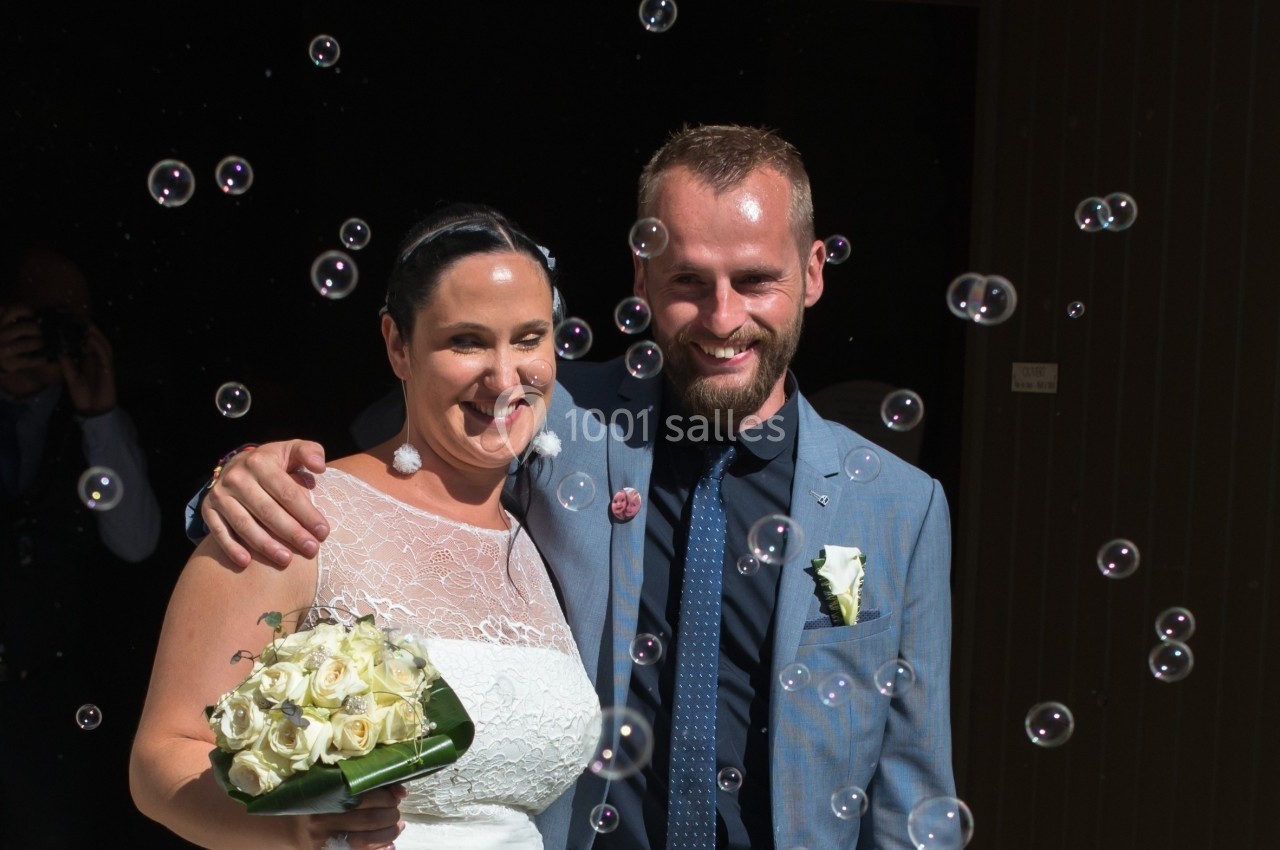 Un couple souriant, vêtu en tenue de mariage, entouré de bulles de savon, à l'extérieur d'un bâtiment sombre.