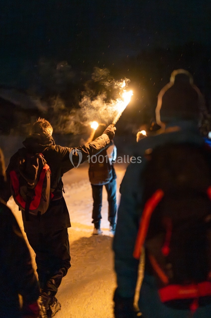 Un groupe de personnes marche de nuit sur un chemin enneigé, éclairé par des torches enflammées.