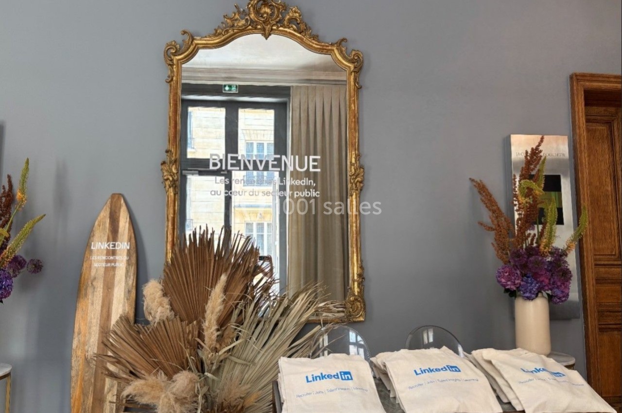 Un miroir doré sur un mur gris reflète une fenêtre, avec des sacs et des décorations florales sur une table en dessous.