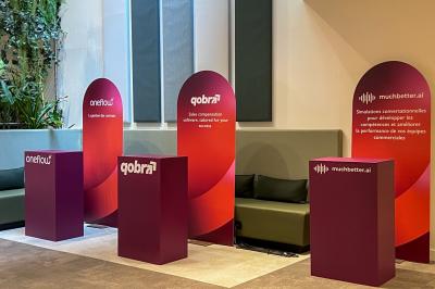 Trois stands promotionnels avec panneaux rouges et logos d'entreprises, disposés devant des banquettes dans un espace…