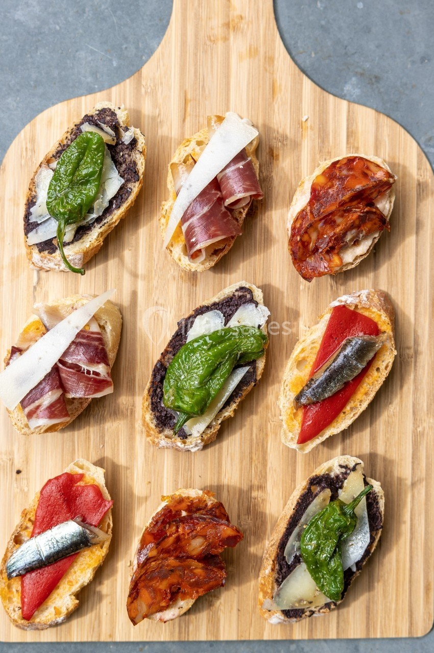 Assortiment de tapas sur une planche en bois, garnis de légumes, charcuterie, poissons et fromage.