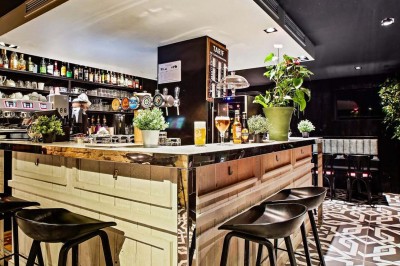 Salle de restaurant avec tables alignées, chaises noires, bouteilles de vin, plantes décoratives et murs en bois.