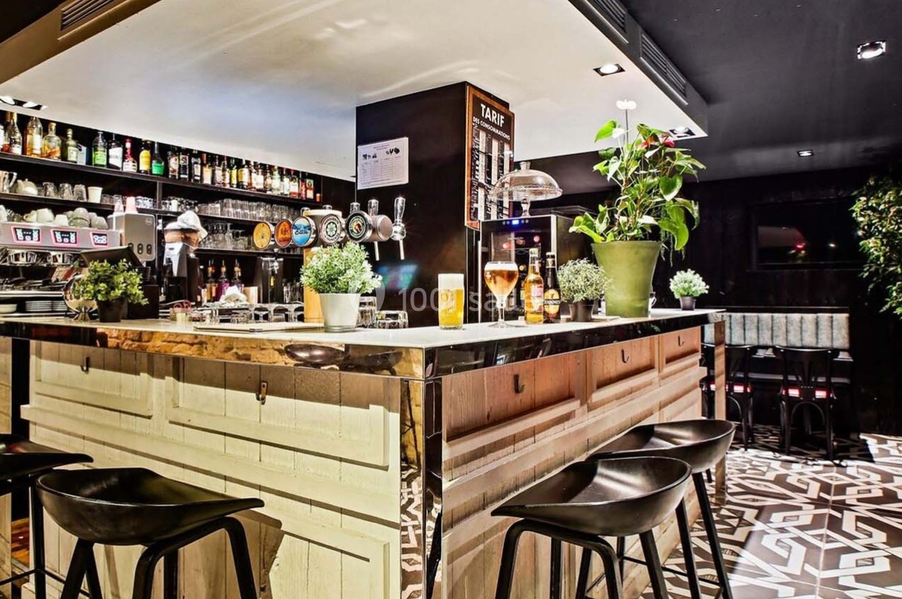 Comptoir de bar moderne avec tabourets noirs, plantes décoratives et étagères remplies de bouteilles en arrière-plan.