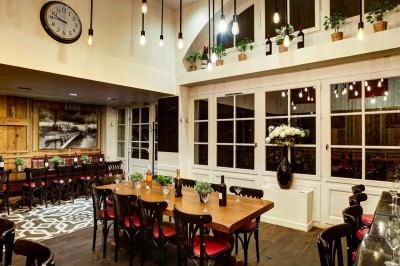 Salle de restaurant avec tables alignées, chaises noires, bouteilles de vin, plantes décoratives et murs en bois.