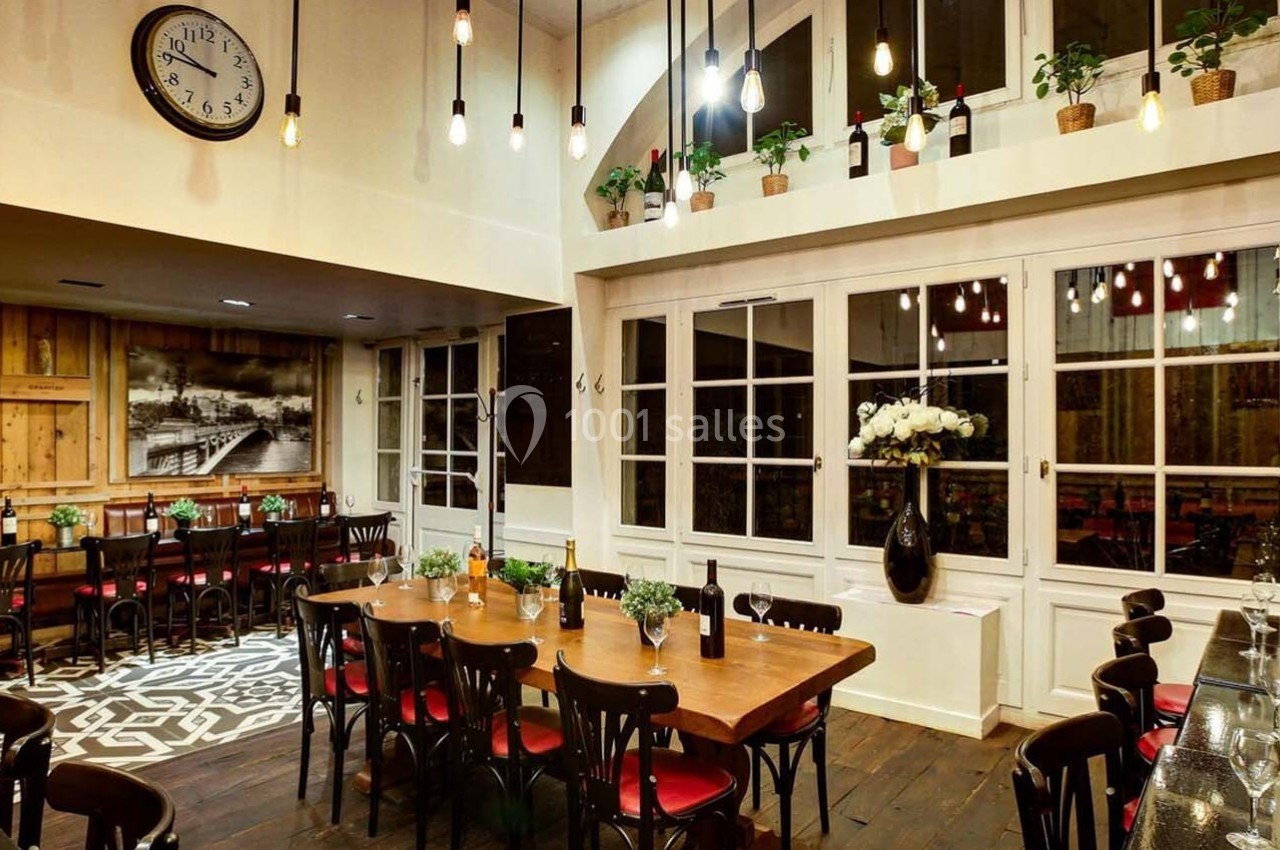 Salle de restaurant chaleureuse avec grandes tables en bois, chaises noires, plantes suspendues et éclairage tamisé.