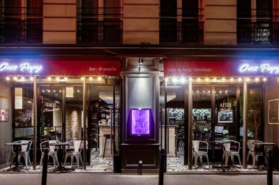 Salle de restaurant avec tables alignées, chaises noires, bouteilles de vin, plantes décoratives et murs en bois.