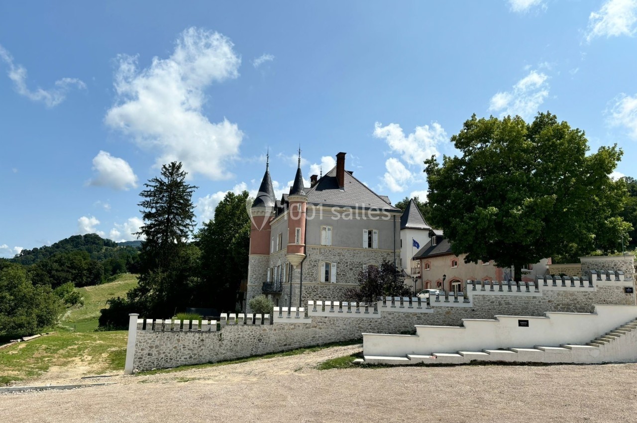 Château entouré de verdure avec ciel bleu, arbres et collines en arrière-plan.
