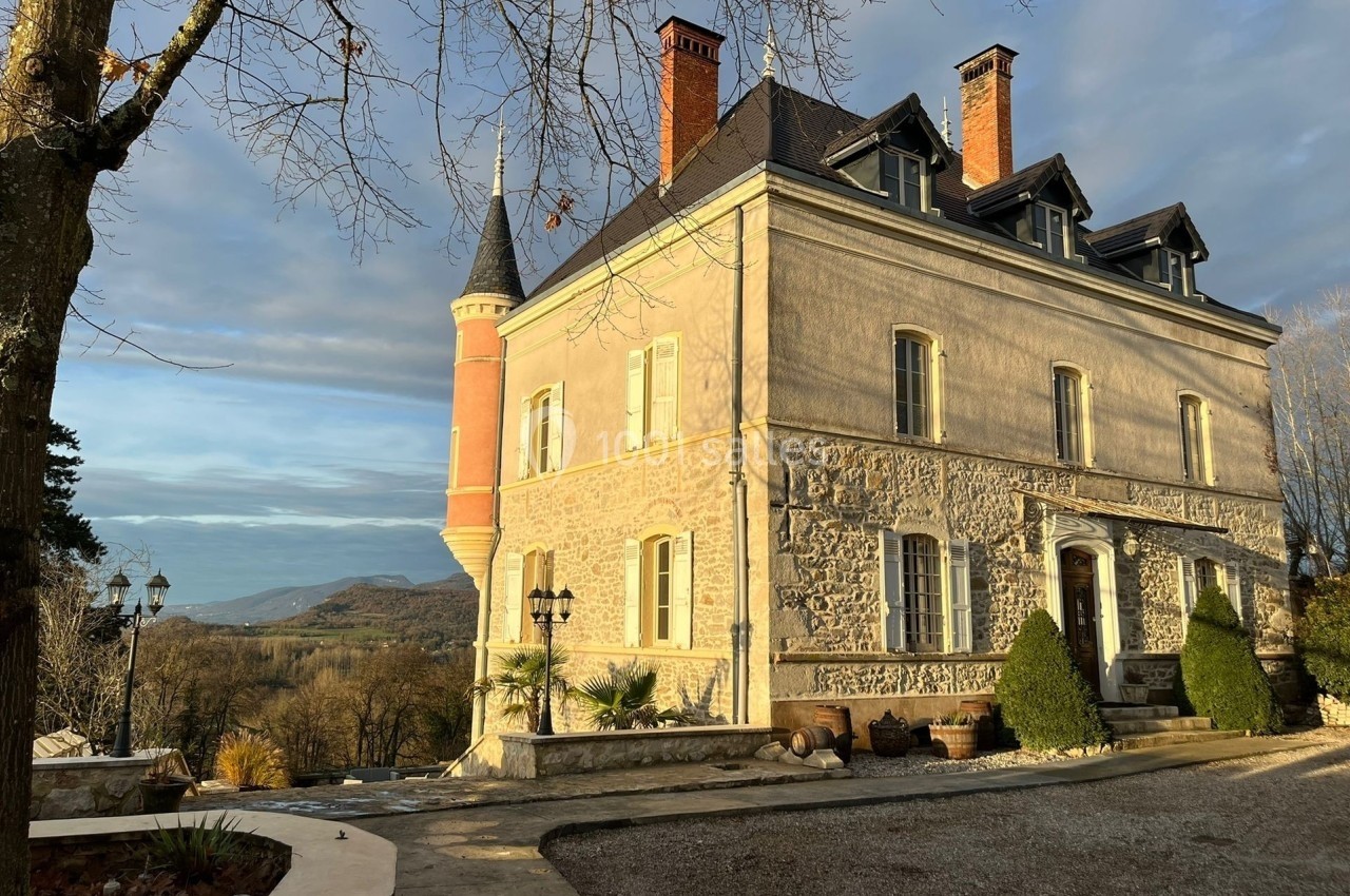 Façade d'un château en pierre avec une tour, entouré d'arbres et offrant une vue sur un paysage vallonné.