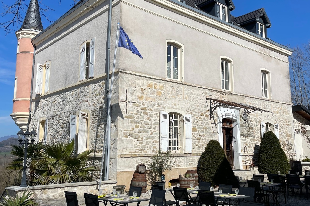 Façade d'un bâtiment ancien avec terrasse aménagée, tables et chaises, sous un ciel bleu dégagé.