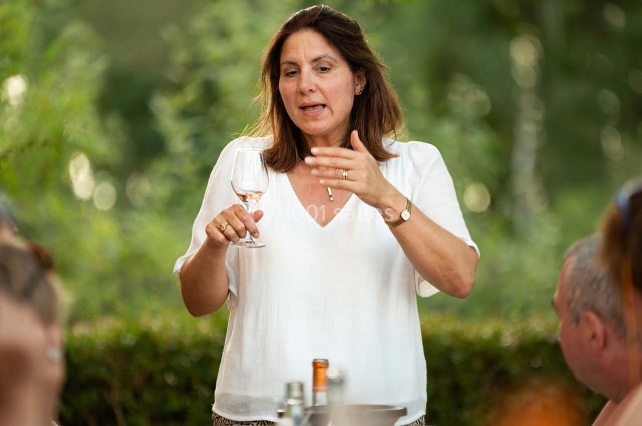 Une femme debout, tenant un verre de vin, parle à un groupe de personnes en extérieur, entourée de verdure.