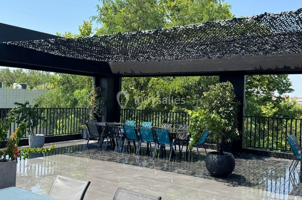 Terrasse extérieure avec tables, chaises bleues et noires, pergola en filet d'ombrage et végétation environnante.