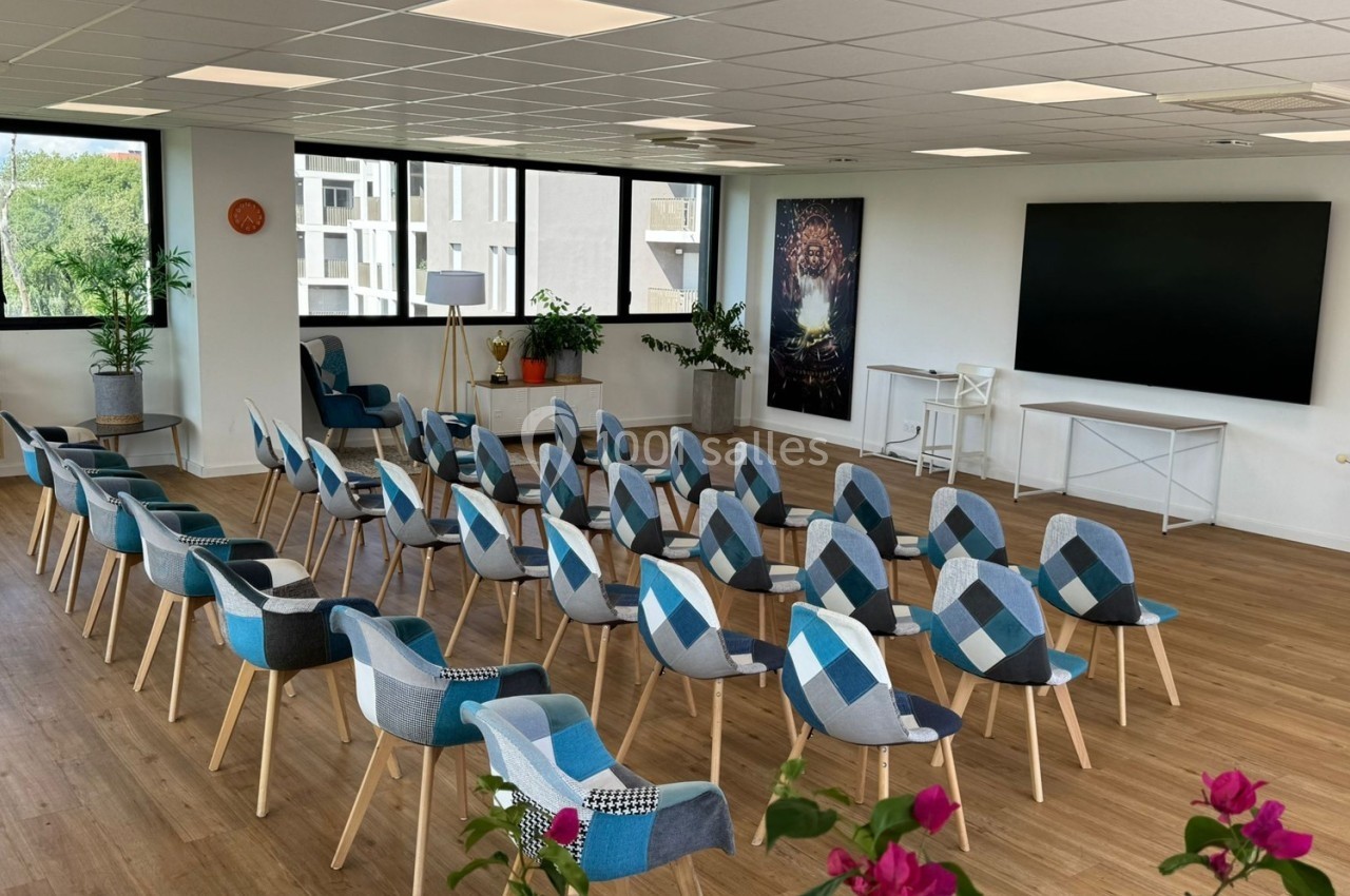 Salle lumineuse avec des rangées de chaises colorées, un écran noir au mur et des plantes en décoration.