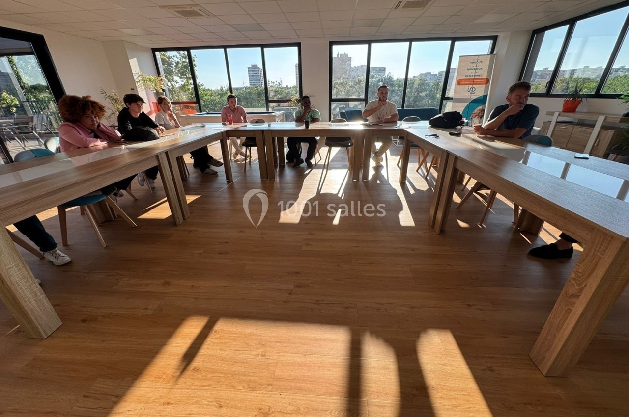 Personnes assises autour de tables en U dans une salle lumineuse avec de grandes fenêtres donnant sur l'extérieur.