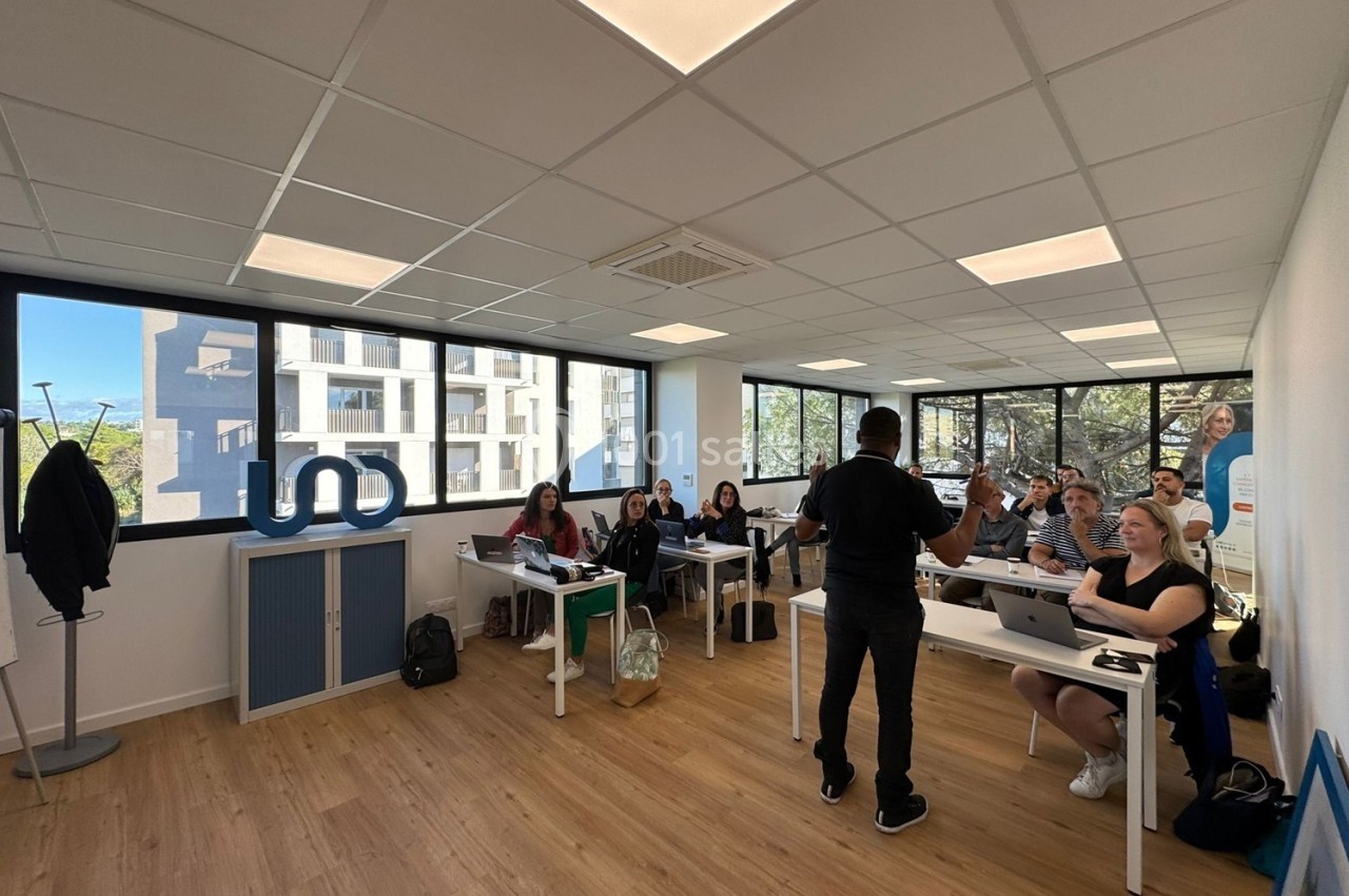Un formateur s'adresse à un groupe d'apprenants dans une salle lumineuse avec des fenêtres donnant sur l'extérieur.