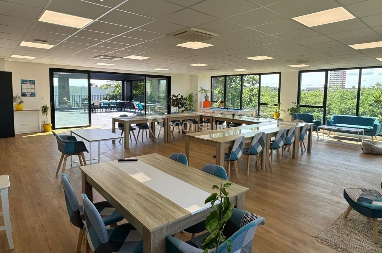 Salle lumineuse avec grandes baies vitrées, tables en bois clair, chaises bleues et espace détente avec canapés.