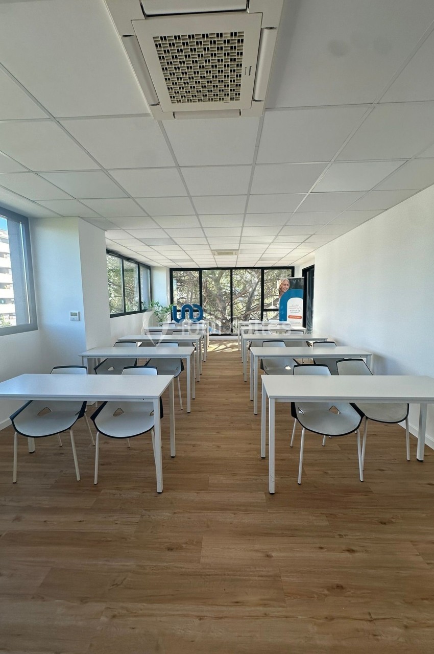 Salle de formation lumineuse avec des tables blanches, des chaises noires et de grandes fenêtres donnant sur l'extérieur.