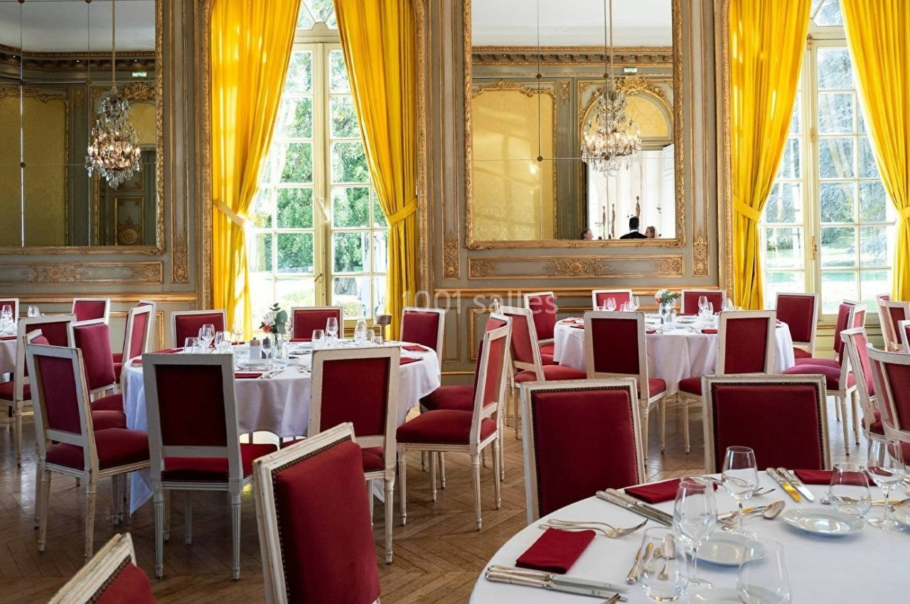 Salle de réception élégante avec des tables dressées, des chaises rouges et des rideaux jaunes encadrant de grandes fenêtres.
