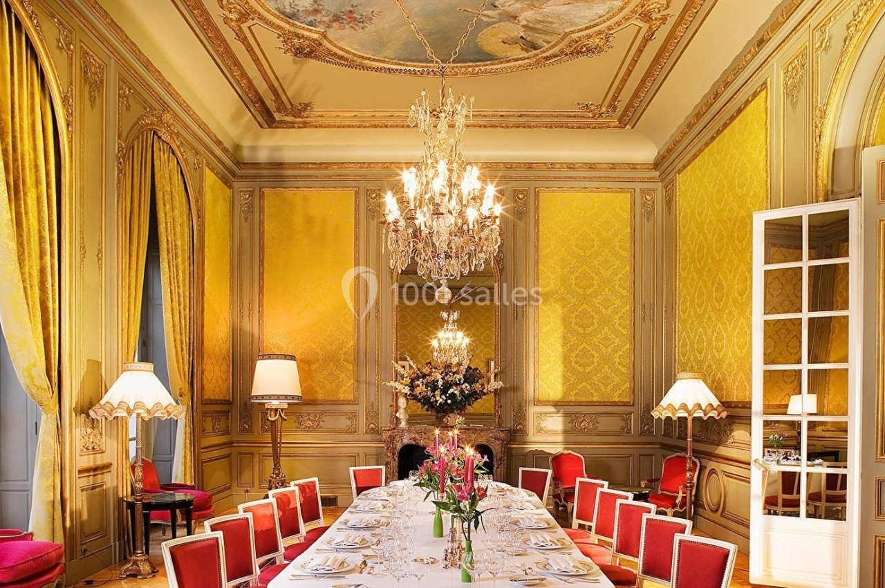 Salle à manger élégante avec un long table dressée, lustres en cristal, murs dorés et plafonds ornés de fresques.