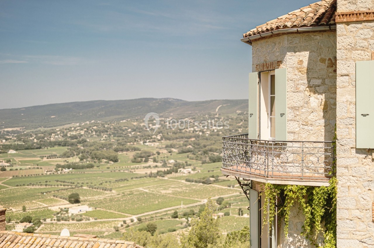 Façade en pierre d'une maison avec balcon, vue sur une vallée verdoyante et des collines sous un ciel dégagé.