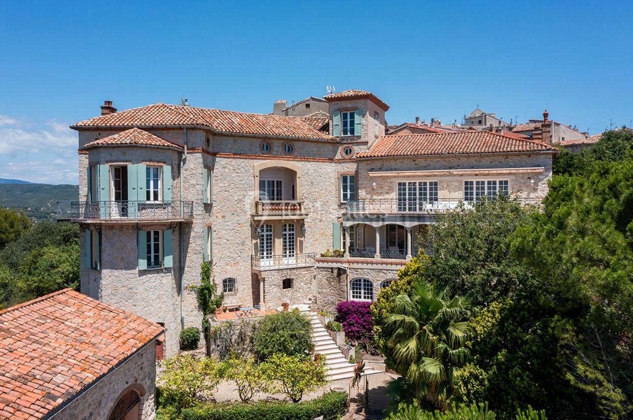 Grande maison en pierre avec toit en tuiles, entourée de végétation et située sous un ciel bleu clair.