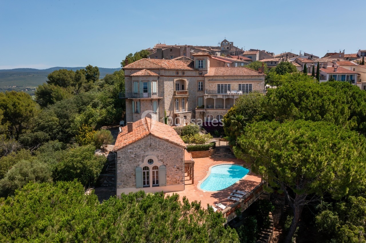 Vue aérienne d'une maison en pierre avec piscine, entourée de végétation et située près d'un village méditerranéen.